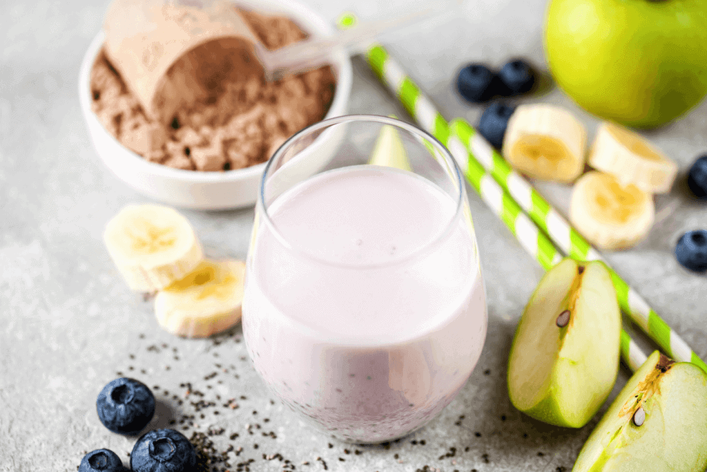 protein shake in clear glass with protein powder in the back and fresh fruits (apples, bananas and blueberries) surrounding