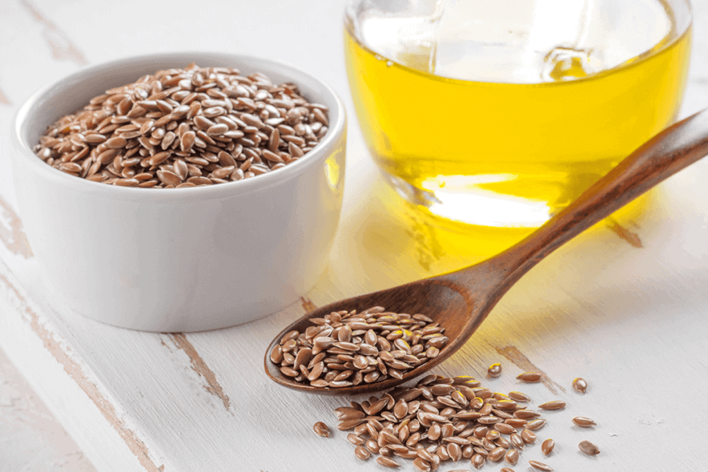 Flaxseeds in a white bowl next to clear jar of flaxseed oil and a wooden spoon of flaxseeds