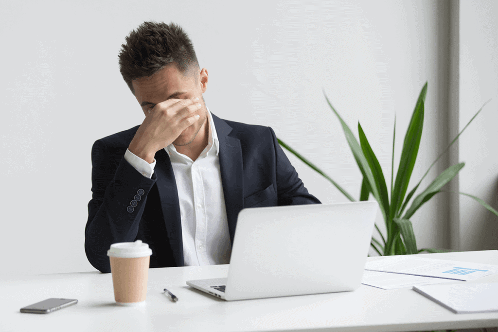 Man sitting at a desk with a laptop and a coffee putting his hand over his eyes as if he's exhausted